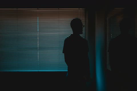 A standing figure in silhouette before blue window blinds